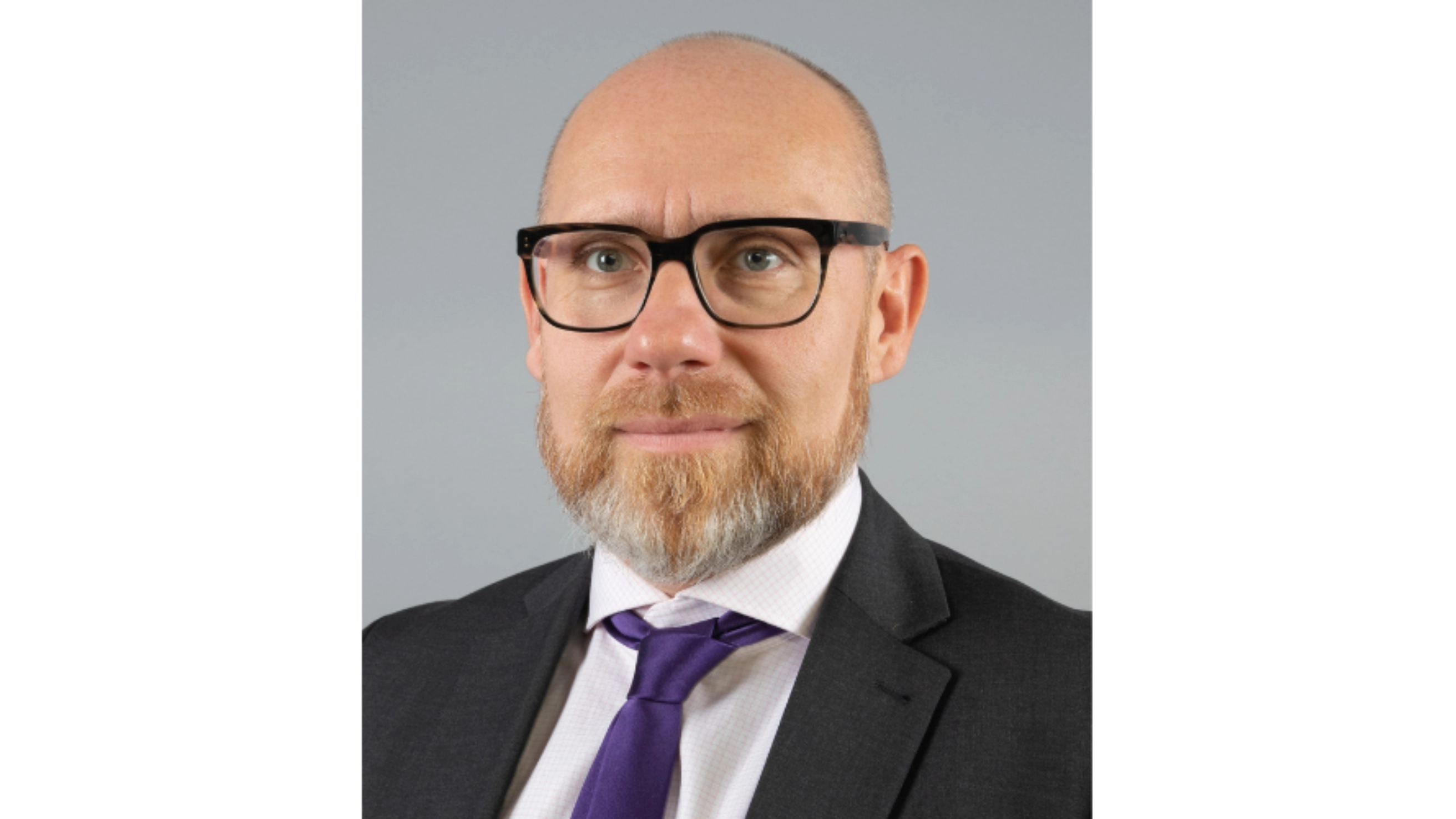 Matthias Galus blickt offen in die Kamera. Er trägt eine Brille und einen eleganten Anzug mit violetter Kravatte.